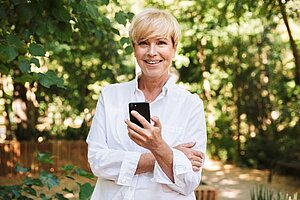 ältere Frau lächelt in die Kamera und hält ein Smartphone in der Hand