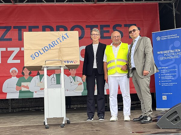 Die KVBW-Vorstände Dr. Doris Reinhardt und Dr. Karsten Braun drücken ihre Solidarität für den Ärzteprotest in Form eines Geschenks aus, das sie stellvertretend Dr. Michael Eckstein überreichen. Die KVBW-Vorstände Dr. Doris Reinhardt und Dr. Karsten Braun drücken ihre Solidarität für den Ärzteprotest in Form eines Geschenks aus, das sie stellvertretend Dr. Michael Eckstein überreichen.