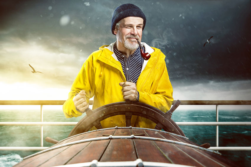alter Kapitän mit gelbem Regenmantel und Pfeife am Steuer eines Schiffs